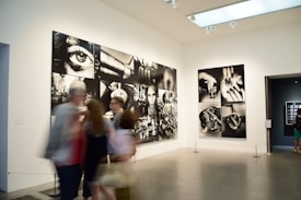 A gallery with modern art photographs displayed on white walls, featuring large black and white images with various abstract and surreal elements. Several people are walking past, slightly blurred, indicating motion.