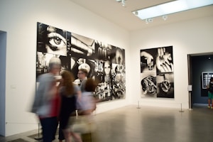 A gallery with modern art photographs displayed on white walls, featuring large black and white images with various abstract and surreal elements. Several people are walking past, slightly blurred, indicating motion.
