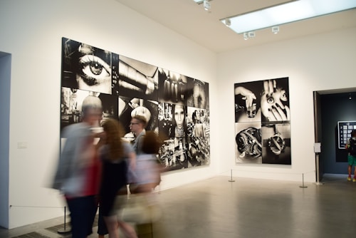 A gallery with modern art photographs displayed on white walls, featuring large black and white images with various abstract and surreal elements. Several people are walking past, slightly blurred, indicating motion.