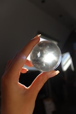 A hand with long nails holding a clear crystal ball, with light shining through it and creating a bright spot. The background appears dark and shadowy, with some structures visible in the light.