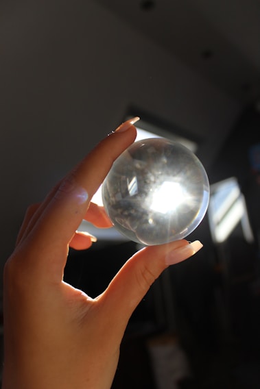A hand with long nails holding a clear crystal ball, with light shining through it and creating a bright spot. The background appears dark and shadowy, with some structures visible in the light.