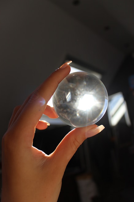 A hand with long nails holding a clear crystal ball, with light shining through it and creating a bright spot. The background appears dark and shadowy, with some structures visible in the light.