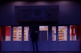 A person stands in front of a small convenience store with brightly lit interior and stocked shelves visible through large glass doors. The exterior is dimly lit, creating a stark contrast with the inside. The scene is likely taking place after dusk or before dawn.