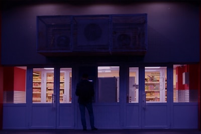 A person stands in front of a small convenience store with brightly lit interior and stocked shelves visible through large glass doors. The exterior is dimly lit, creating a stark contrast with the inside. The scene is likely taking place after dusk or before dawn.