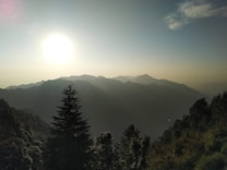 The image portrays a breathtaking mountain landscape during sunrise or sunset, with rolling hills and a bright sun casting a warm glow across the sky. Silhouettes of trees are visible in the foreground, creating a serene and peaceful atmosphere.