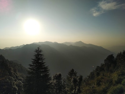 The image portrays a breathtaking mountain landscape during sunrise or sunset, with rolling hills and a bright sun casting a warm glow across the sky. Silhouettes of trees are visible in the foreground, creating a serene and peaceful atmosphere.