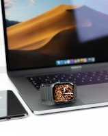 A smartwatch with a unique art design on the screen is placed on a laptop's trackpad. The laptop is open, displaying a wallpaper of sand dunes. Next to it is a smartphone, likely an iPhone, with a reflective surface.