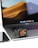 A smartwatch with a unique art design on the screen is placed on a laptop's trackpad. The laptop is open, displaying a wallpaper of sand dunes. Next to it is a smartphone, likely an iPhone, with a reflective surface.