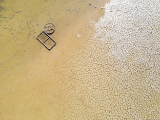 Aerial view of a cracked, dry earth surface with a circular and rectangular metallic object lying on it. The ground displays a pattern of fissured, parched soil, suggesting arid conditions.