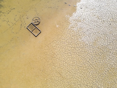 Aerial view of a cracked, dry earth surface with a circular and rectangular metallic object lying on it. The ground displays a pattern of fissured, parched soil, suggesting arid conditions.