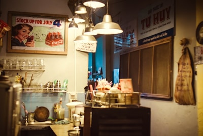 A vintage kitchen or café interior featuring various kitchenware and utensils neatly arranged on a counter. A retro-style advertisement poster and a 'Thank You, Come Again' sign are visible on the walls, alongside several industrial-style hanging lamps providing soft, warm lighting.