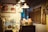 A vintage kitchen or café interior featuring various kitchenware and utensils neatly arranged on a counter. A retro-style advertisement poster and a 'Thank You, Come Again' sign are visible on the walls, alongside several industrial-style hanging lamps providing soft, warm lighting.