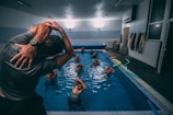 A group of individuals is engaged in a water aerobics class in an indoor swimming pool. The instructor, wearing a wristwatch, is demonstrating a stretch with their arms lifted over their head. Several participants in the pool are following the instructor’s lead, with their arms similarly raised. Colorful pool noodles are scattered along the poolside. Various towels hang on the wall of the pool area.