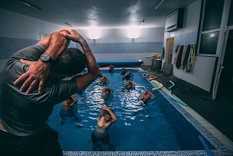 A group of individuals is engaged in a water aerobics class in an indoor swimming pool. The instructor, wearing a wristwatch, is demonstrating a stretch with their arms lifted over their head. Several participants in the pool are following the instructor’s lead, with their arms similarly raised. Colorful pool noodles are scattered along the poolside. Various towels hang on the wall of the pool area.