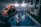 A group of individuals is engaged in a water aerobics class in an indoor swimming pool. The instructor, wearing a wristwatch, is demonstrating a stretch with their arms lifted over their head. Several participants in the pool are following the instructor’s lead, with their arms similarly raised. Colorful pool noodles are scattered along the poolside. Various towels hang on the wall of the pool area.