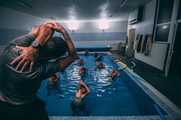A group of individuals is engaged in a water aerobics class in an indoor swimming pool. The instructor, wearing a wristwatch, is demonstrating a stretch with their arms lifted over their head. Several participants in the pool are following the instructor’s lead, with their arms similarly raised. Colorful pool noodles are scattered along the poolside. Various towels hang on the wall of the pool area.