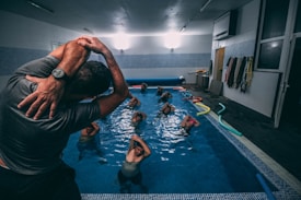A group of individuals is engaged in a water aerobics class in an indoor swimming pool. The instructor, wearing a wristwatch, is demonstrating a stretch with their arms lifted over their head. Several participants in the pool are following the instructor’s lead, with their arms similarly raised. Colorful pool noodles are scattered along the poolside. Various towels hang on the wall of the pool area.