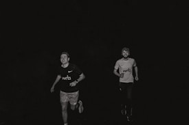 Two people are running outdoors at night. Both are wearing athletic clothing with visible logos on their shirts. The image is in black and white, highlighting the contrast between their clothing and the dark background.