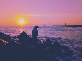 A stylish angler sporting a fish gear hat, standing on a rocky shoreline with a sunset backdrop.