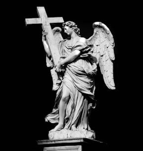 A majestic stone sculpture of an angel holding a large cross. The angel is depicted with detailed feathered wings and flowing drapery, conveying a sense of grace and elegance. The sculpture is set against a dark background, highlighting the intricate features of the artwork.