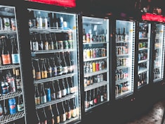 A row of refrigerated display cases filled with a variety of bottled and canned beverages. The shelves are neatly organized with diverse labels and colors visible on the containers. The refrigeration units have illuminated interiors, enhancing the vibrant colors of the labels.