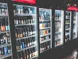 A row of refrigerated display cases filled with a variety of bottled and canned beverages. The shelves are neatly organized with diverse labels and colors visible on the containers. The refrigeration units have illuminated interiors, enhancing the vibrant colors of the labels.