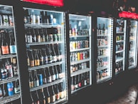 A row of refrigerated display cases filled with a variety of bottled and canned beverages. The shelves are neatly organized with diverse labels and colors visible on the containers. The refrigeration units have illuminated interiors, enhancing the vibrant colors of the labels.