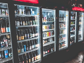 A row of refrigerated display cases filled with a variety of bottled and canned beverages. The shelves are neatly organized with diverse labels and colors visible on the containers. The refrigeration units have illuminated interiors, enhancing the vibrant colors of the labels.