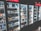 A row of refrigerated display cases filled with a variety of bottled and canned beverages. The shelves are neatly organized with diverse labels and colors visible on the containers. The refrigeration units have illuminated interiors, enhancing the vibrant colors of the labels.