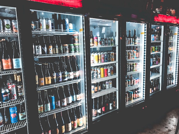 A row of refrigerated display cases filled with a variety of bottled and canned beverages. The shelves are neatly organized with diverse labels and colors visible on the containers. The refrigeration units have illuminated interiors, enhancing the vibrant colors of the labels.