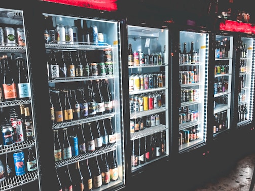 A row of refrigerated display cases filled with a variety of bottled and canned beverages. The shelves are neatly organized with diverse labels and colors visible on the containers. The refrigeration units have illuminated interiors, enhancing the vibrant colors of the labels.