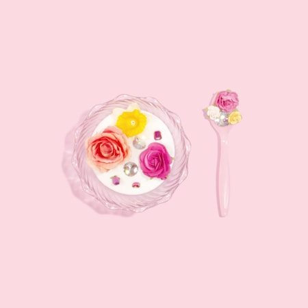 A delicate arrangement featuring a glass bowl with a creamy white substance topped with colorful artificial flowers and small, shiny gemstones. Beside the bowl, there is a pink spoon also adorned with similar flowers and gems, all set against a pastel pink background.