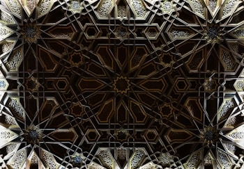 Intricate geometric patterns form a symmetrical design, featuring interlacing shapes that create an elaborate mosaic. The design includes star shapes and hexagons, with fine detailing and a sense of depth.