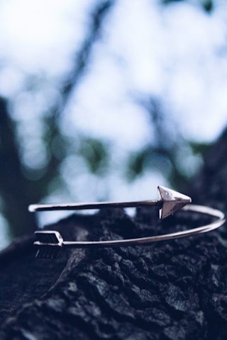 A metal bracelet with an arrow design resting on a textured, dark surface. The background features a blurred view of a tree with bokeh effect, highlighting the natural setting.