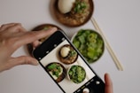 A hand is holding a smartphone, capturing a photo of a meal with several dishes. The dishes include a bowl of salad with greens, a plate of food containing a bun or dumpling, and noodles garnished with fresh herbs. Chopsticks are placed alongside the bowls on the table.