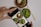 A hand is holding a smartphone, capturing a photo of a meal with several dishes. The dishes include a bowl of salad with greens, a plate of food containing a bun or dumpling, and noodles garnished with fresh herbs. Chopsticks are placed alongside the bowls on the table.