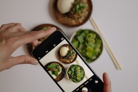 A hand is holding a smartphone, capturing a photo of a meal with several dishes. The dishes include a bowl of salad with greens, a plate of food containing a bun or dumpling, and noodles garnished with fresh herbs. Chopsticks are placed alongside the bowls on the table.