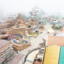 An amusement park under construction featuring winding roller coaster tracks and artificial rocky landscapes with a misty atmosphere. Buildings and scaffolding are visible, along with pathways and pools of water.