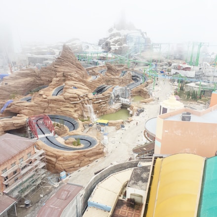 An amusement park under construction featuring winding roller coaster tracks and artificial rocky landscapes with a misty atmosphere. Buildings and scaffolding are visible, along with pathways and pools of water.