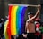 A person wearing a leather outfit is holding a rainbow flag, commonly associated with LGBTQ+ pride, in a public gathering. The crowd around includes individuals of diverse appearances, some of whom are also dressed in colorful attire.