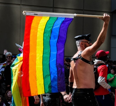 A person wearing a leather outfit is holding a rainbow flag, commonly associated with LGBTQ+ pride, in a public gathering. The crowd around includes individuals of diverse appearances, some of whom are also dressed in colorful attire.