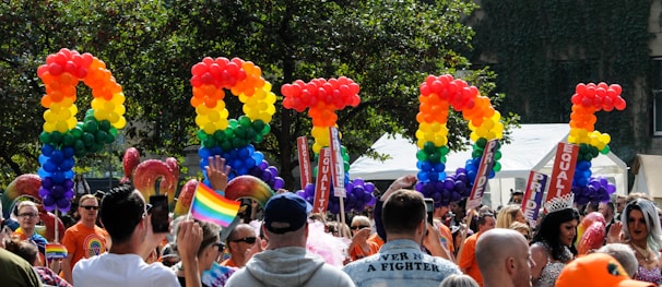 A vibrant and lively celebration featuring a diverse crowd of people. Brightly colored balloons spell out the word 'PRIDE' against a backdrop of trees. Participants are waving rainbow flags and wearing colorful outfits. Signs with the words 'EQUALITY' are visible among the crowd.