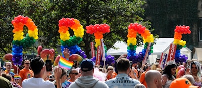 A vibrant and lively celebration featuring a diverse crowd of people. Brightly colored balloons spell out the word 'PRIDE' against a backdrop of trees. Participants are waving rainbow flags and wearing colorful outfits. Signs with the words 'EQUALITY' are visible among the crowd.
