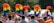 A vibrant and lively celebration featuring a diverse crowd of people. Brightly colored balloons spell out the word 'PRIDE' against a backdrop of trees. Participants are waving rainbow flags and wearing colorful outfits. Signs with the words 'EQUALITY' are visible among the crowd.