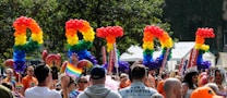 A vibrant and lively celebration featuring a diverse crowd of people. Brightly colored balloons spell out the word 'PRIDE' against a backdrop of trees. Participants are waving rainbow flags and wearing colorful outfits. Signs with the words 'EQUALITY' are visible among the crowd.