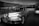 A classic retro car is prominently displayed in a showroom with the word 'REINVENTION' in large letters on the wall behind it. The car exhibits a streamlined design and is positioned towards the front, with another car partially visible in the background. The black and white setting adds a timeless quality to the scene.