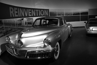 A classic retro car is prominently displayed in a showroom with the word 'REINVENTION' in large letters on the wall behind it. The car exhibits a streamlined design and is positioned towards the front, with another car partially visible in the background. The black and white setting adds a timeless quality to the scene.