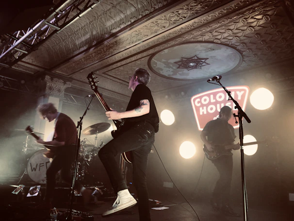 A band performs energetically on stage with illuminated circular lights in the background. The ceiling features ornate decorations, and there is a prominent sign reading 'Colony House'. The musicians are engaged with guitars and a drum set is visible.