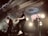 A band performs energetically on stage with illuminated circular lights in the background. The ceiling features ornate decorations, and there is a prominent sign reading 'Colony House'. The musicians are engaged with guitars and a drum set is visible.