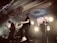 A band performs energetically on stage with illuminated circular lights in the background. The ceiling features ornate decorations, and there is a prominent sign reading 'Colony House'. The musicians are engaged with guitars and a drum set is visible.
